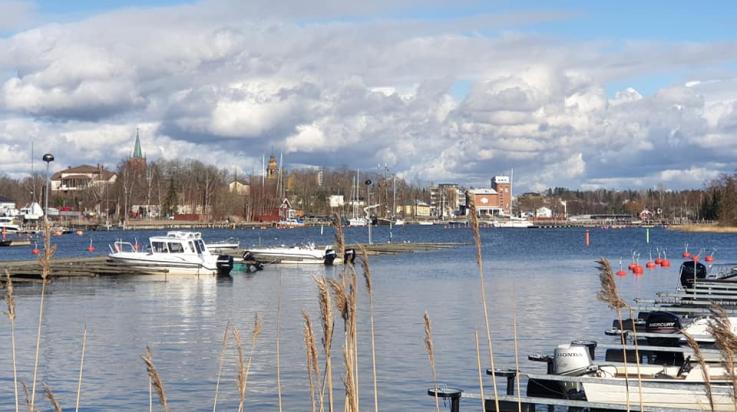 Kaunis, huhtikuinen kuva Kaupunginlahden alkupäästä, jossa näkyy edessä kaisloja, niiden takana laitureissaan olevia veneitä, ja taaempana näkyy kaupunki.